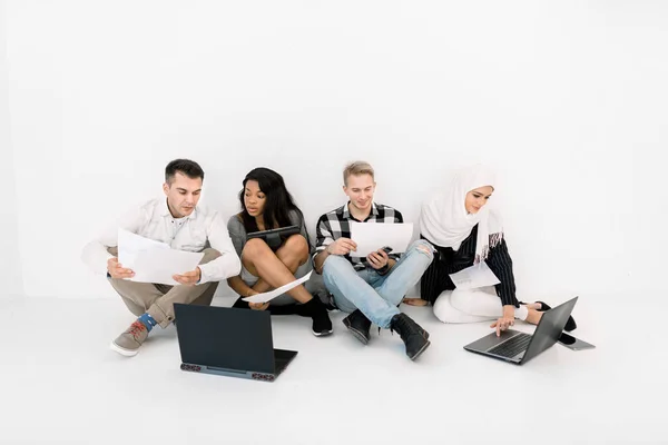 Grupo de cuatro jóvenes creativos multiétnicos que planean un nuevo proyecto, hablando entre sí, usando un ordenador portátil y una tableta, mientras están sentados en el suelo sobre un fondo blanco — Foto de Stock