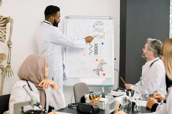 Interracial group of doctors, chemists, scientists having discussion in meeting while young African man telling new information and showing biochemical molecules of substances on the board.