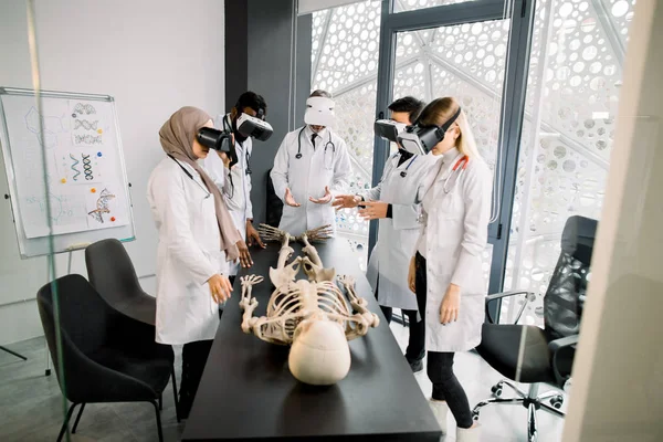 science, technology, virtual reality and people concept. Professor with group of multiethnic medical students using virtual reality goggles to exam human skeleton lying on the table