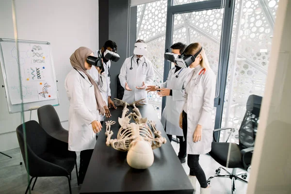 Science, technology, virtual reality and people concept. Professor with group of multiethnic medical students using virtual reality goggles to exam human skeleton lying on the table — Stok fotoğraf
