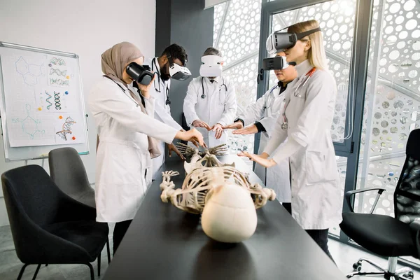 Education, science, vr, medical and health concept. Group of multiethnic doctors, biologists, archeologists, scientists wearing VR goggles study human skeleton in modern laboratory — Stock Photo, Image