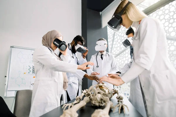Equipe de médicos multiétnicos profissionais, cirurgiões, ortopedistas, estudantes vestindo óculos VR trabalhando com modelo de esqueleto humano em sala de aula moderna. Anatomia virtual, conceito de medicina — Fotografia de Stock