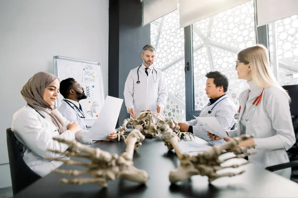 Beau médecin d'âge moyen, professeur d'anatomie, expliquant l'anatomie osseuse à l'aide d'un modèle squelette pour divers étudiants en blouse blanche. Médecine, éducation, concept d'anatomie — Photo