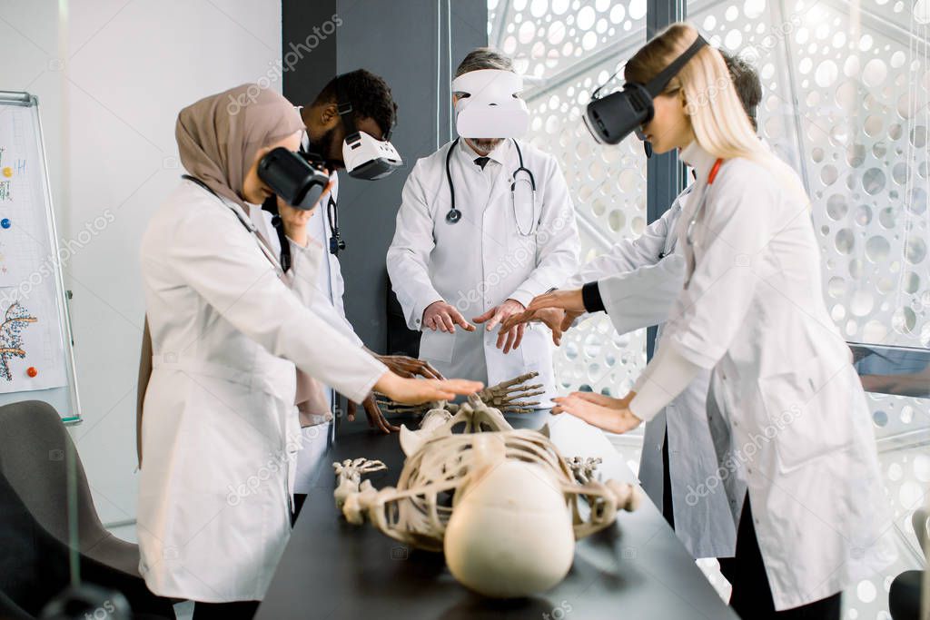 Working atmosphere in modern lab. Concentrated multiethnic professional biologists, doctors, scientists wearing VR glasses working with human skeleton on the table