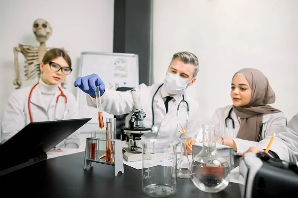 Medicine, genetics, immunology, microbiology concept. Multiethnic group of scientists, Caucasian man and woman, Muslim woman, working at the laboratory with microscope, tubes with liquids and laptop. — Stok fotoğraf
