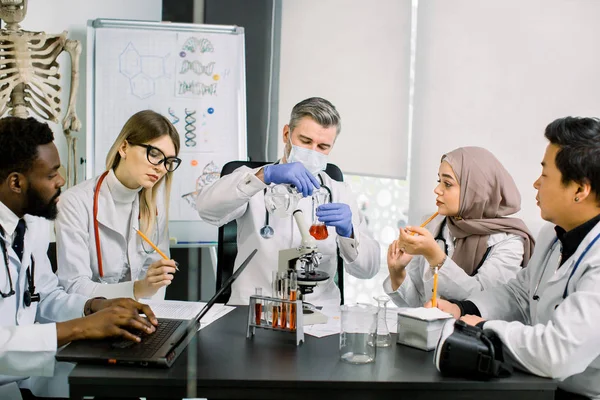 Five young multiethnic doctors scientists, bioengeneers, chemists, working in modern laboratory center. Male scientist holds flask with sample of new substance, African man makes notes on laptop — Stok fotoğraf