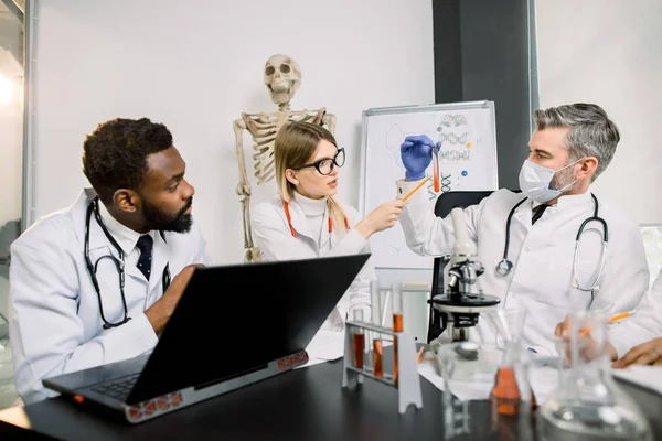 Concept de santé et de biotechnologie. De jeunes scientifiques multiethniques travaillent en laboratoire. Homme masqué montre tube à essai avec une nouvelle substance ou du sang à l'homme africain tapant sur ordinateur portable et femme caucasienne — Photo