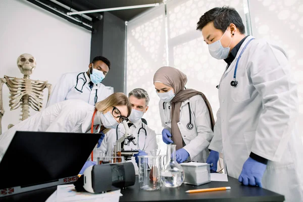 Team of lab workers, veterinarians, bio chemists examining tissues or blood sample using the microscope. Group of multiethnic scietists working with microscope, tubes and flasks, laptop in laboratory