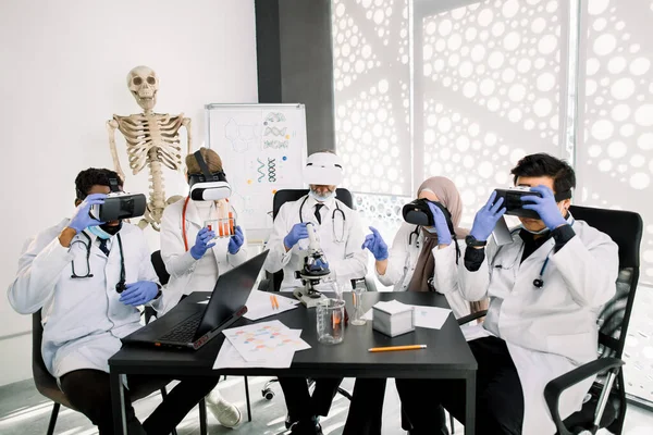 Equipe de farmacologistas multiétnicos vestindo casacos brancos de proteção, luvas e óculos de proteção, trabalhando juntos em novas vacinas ou drogas em laboratório moderno. Laptop, tubos, frascos e papéis sobre a mesa — Fotografia de Stock