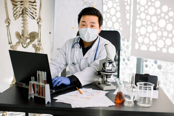 Портрет азіатського вченого, який сидить за столом і робить нотатки на ноутбуці, працюючи з мікроскопом і тестуючи трубки в лабораторії. Медичне, медичне та фармацевтичне дослідження. — стокове фото