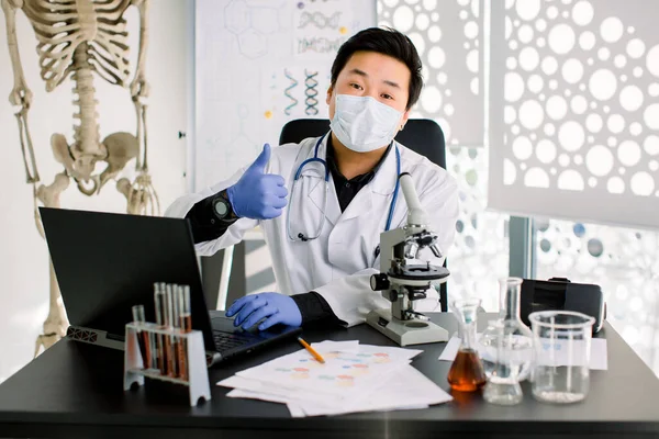 Happy Asian man scientist in white coat, mask and blue gloves, types on laptop the results of research, and shows his thumb up. Synthesis of new drugs, cancer treatment, medicine, biochemistry concept — Stok fotoğraf