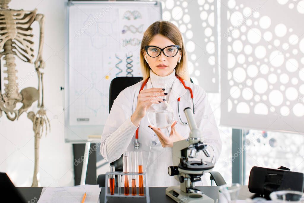 Smart pretty Caucasian young lady scientist shows glass flask with liquid inside, sitting at the table with microscope and test tubes in the laboratory. Human skeleton on background. Science concept