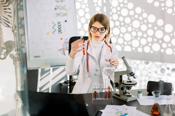 Mulher branca bonita, cientista farmacêutico laboratório, médico, geneticista, examinando amostra de sangue em tubo de teste, trabalhando em laboratório moderno, sentado à mesa com microscópio e laptop — Fotografia de Stock