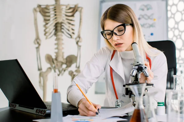 Молода жінка-лікар, біохімік, вчений, дивлячись через мікроскоп в лабораторії, робить нотатки про папери і ноутбук. Світла лабораторна кімната, людський скелет на фоні — стокове фото