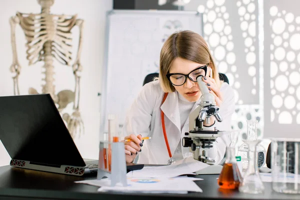 Mikroskopla bakan, laboratuvar ekipmanları ve dizüstü bilgisayarla masada oturan, koruyucu gözlüklü beyaz kadın doktor. Tıbbi analiz, biyoteknoloji, biyoloji. — Stok fotoğraf