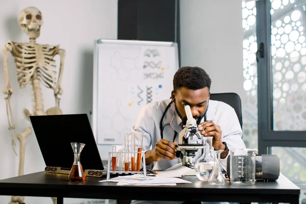 Yakışıklı bir Afrikalı teknisyen, doktor, mikrobiyolog, mikroskopla inceleyen, tıbbi araştırma üzerinde çalışan, modern cam teçhizat ve dizüstü bilgisayarla masada oturan. — Stok fotoğraf