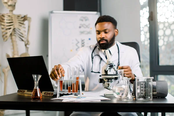 African man scientist researcher sitting at the table in lab and doing dna test, analisys of blood samples or new substances drugs, examining test tubes, working with data on laptop on the table.