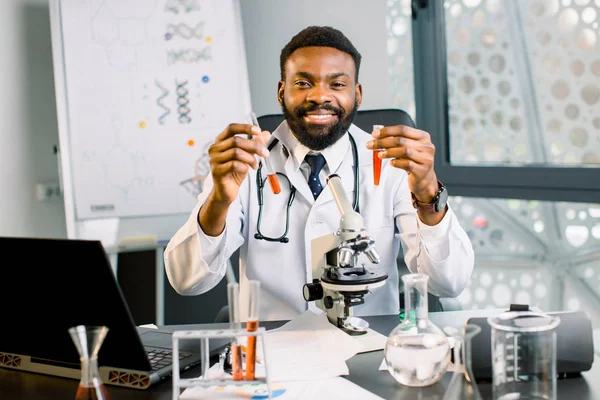Handsome young African male scientist, chemist, doctor wearing lab coat smiling happily holding two test tubes with red liquid, working at the lab. Chemical analysis, medical research concept — Stock Photo, Image