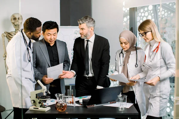 Deux chefs d'entreprise, asiatiques et caucasiens, avec un groupe de chercheurs multiethniques dans un laboratoire moderne, discutant des résultats d'un projet scientifique ou d'une nouvelle start-up — Photo