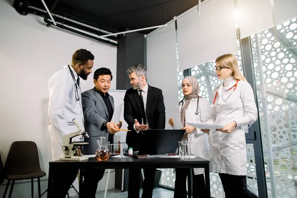 Przystojny dojrzały biznesmen i jego azjatycki partner rozmawiają i spotykają się z grupą trzech wielonarodowych młodych lekarzy lub naukowców stojąc w nowoczesnym biurze w klinice — Zdjęcie stockowe