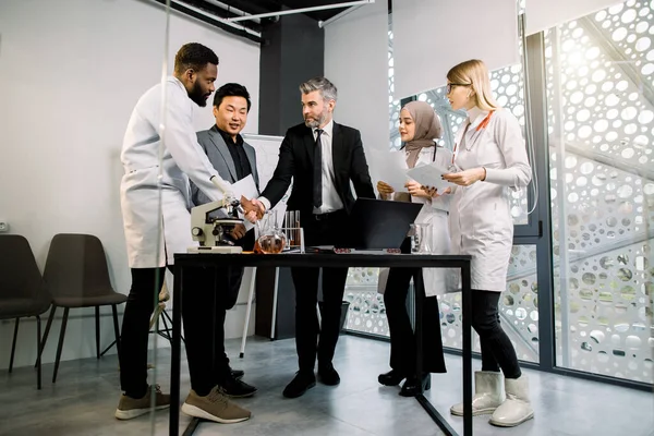 Grupo inter-racial de homens de negócios e médicos cientistas reunião de equipe em laboratório moderno ou escritório, de pé à mesa com laptop, microscópio e óculos. Handshaking de homens caucasianos e africanos — Fotografia de Stock