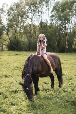 Güzel, gülümseyen beyaz, uzun saçlı abd boho aksesuarları olan küçük bir kız yazın arazide ata biniyor. At binme, çocukluk, kırsal yaşam tarzı.