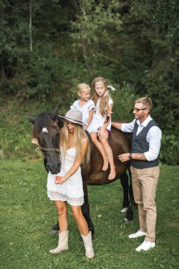 Mutlu aile, baba, anne ve iki çocuk ata binen kızlar yeşil çayırlarda dinleniyor. Aile, şık boho, çiftlik kıyafetleri, açık havada at yürüyüşü.