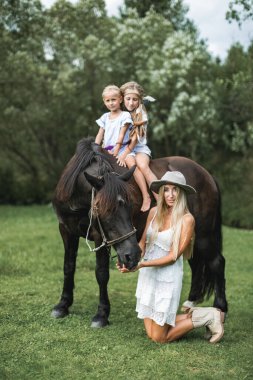Mutlu bir aile, iki çocuklu bir anne ata binen sevimli kızlar. Beyaz elbiseli, şapkalı ve kovboy çizmeli anne dizlerinin üstünde duruyor ve atı besliyor. Doğada aile zamanı, ata binme.
