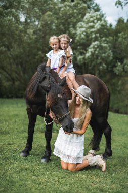 Çekici bir aile, anne ve iki kız şık günlük boho kıyafetleri giyiyorlar, kahverengi bir atın üzerinde çayırda yürüyorlar. Güzel, genç bir anne at besliyor ve iki çocuk sürüyor.