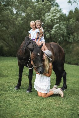 Çekici bir aile, anne ve iki kız şık günlük boho kıyafetleri giyiyorlar, kahverengi bir atın üzerinde çayırda yürüyorlar. Güzel genç anne ve atlarıyla iki çocuk..