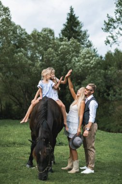 At çiftliğinde at sırtında yürüyen mutlu bir aile. Genç mutlu aile kırsal alanda eğleniyor, iki kız ata biniyor, anne ve baba gülümsüyor ve kızlarına beşlik çakıyor.