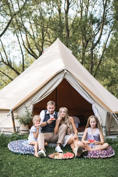 Šťastná rodina nosí kovbojské boho oblečení, otec, matka a dvě dcery sedí na polštářích na trávě a jedí meloun. Piknik. Summer. Big tipi wigwam na pozadí — Stock fotografie