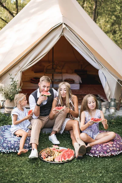 Kavkazská rodina má piknik a kempování v lese, sedí před velkým vigvamovým stanem, relaxují a cítí se šťastní, jedí spolu sladký meloun. Rodinná aktivita, příroda a sluneční světlo — Stock fotografie