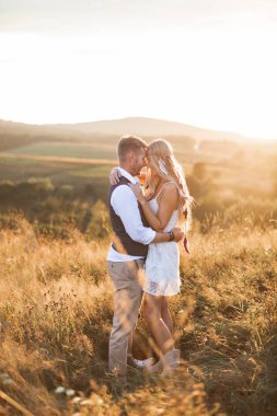 Günbatımında tarlada, dağların arka planında poz veren şık boho kıyafetleri içindeki kadın ve erkek. Havalı hippi çift sarılıyor.