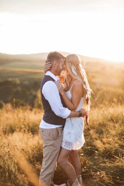 Kırsal, boho ve insanlar konsepti. Saçında tüylü aksesuarları olan bir kadın ve tarlada poz veren şık giyinmiş yakışıklı bir adam.