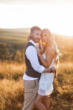 beautiful couple dressed in boho style embracing each other in the sunny summer field. Handsome man in casual wear and woman in white dress and feathers in hair walking in the field at sunset