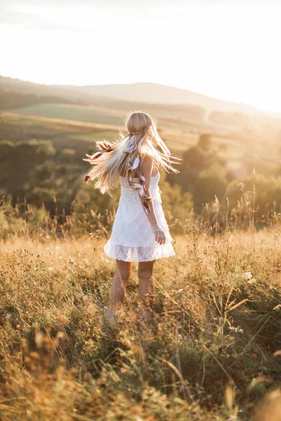 Menina jovem hipster em vestido branco e penas de cabelo em pé ao ar livre, no campo de verão nas montanhas. Estilo hippie Boho — Fotografia de Stock