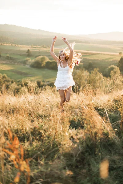 Menina boho muito jovem em uma colina com fundo de campo de verão. Mulher bonita em vestido branco em estilo boho com uma coroa de penas no cabelo, pulando e andando no verão no campo . — Fotografia de Stock