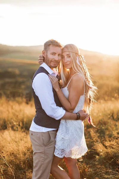 Belo casal vestido em estilo boho abraçando uns aos outros no campo de verão ensolarado. Bonito homem em casual desgaste e mulher em vestido branco e penas no cabelo andando no campo ao pôr do sol — Fotografia de Stock