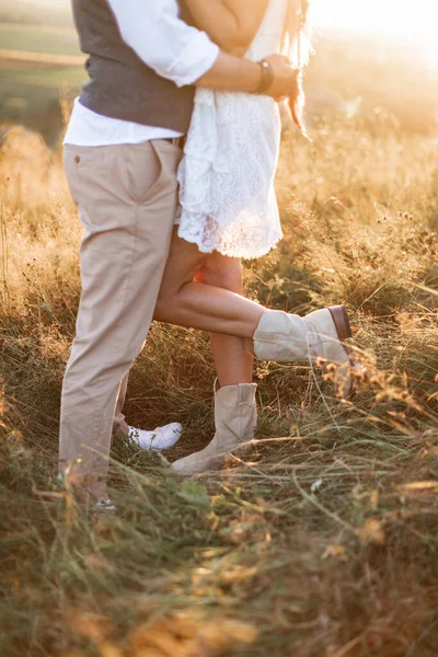 Ausgeschnittene Bild der Beine von stilvollen rustikalen Paar, Boho Frau in weißem Kleid und Cowboystiefel und Mann in Hemd und Hose posiert in sonnigem Feld, umarmen einander — Stockfoto