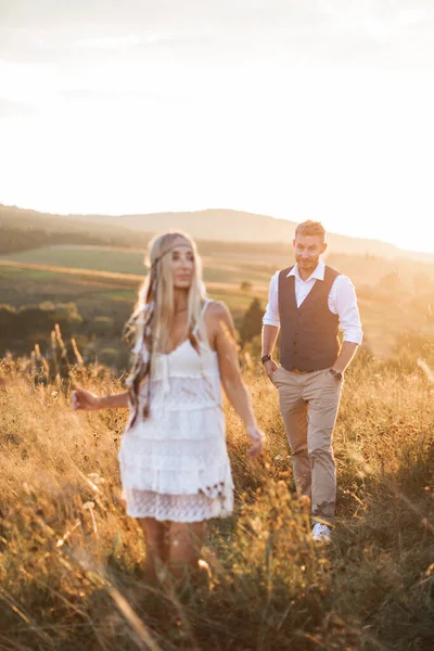 Élégant bel homme marchant vers sa femme heureuse en vêtements boho dans la lumière du soleil, moment romantique en plein air dans le champ de coucher de soleil d'été. Concentre-toi sur l'homme — Photo