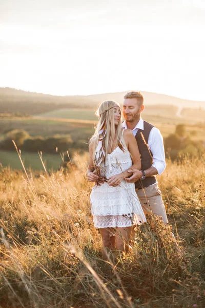 Un bel homme étreint sa femme debout dans le champ avec de l'herbe de paille et des fleurs au coucher du soleil d'été, à l'extérieur. Mariage, histoire d'amour dans le style rustique boho — Photo