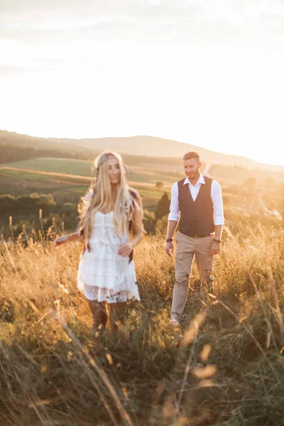 Homem elegante bonito andando para sua mulher feliz em roupas boho na luz do sol, momento romântico ao ar livre no campo de pôr do sol de verão. Concentre-se no homem — Fotografia de Stock