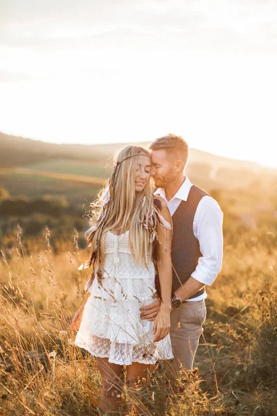 Beautiful couple in love embracing, at the sunset in summer field. Girl in white summer dress and feathers in hair, man in stylish casual suit. Summer vacation concept, boho couple — Zdjęcie stockowe