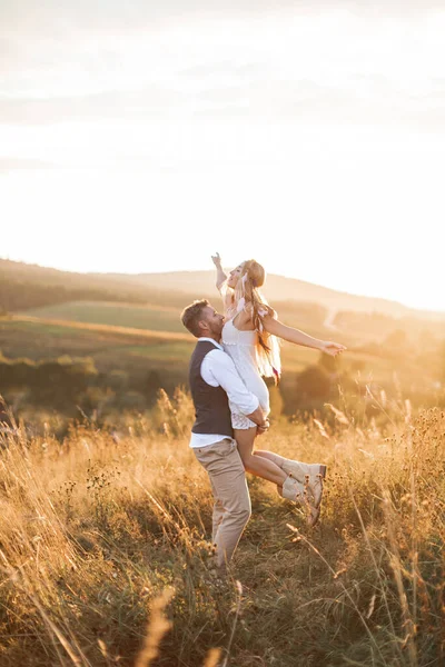 Casal jovem vestindo roupas estilo hipster boho indie, no amor andando no campo, o homem mantém a mulher a mãos, a mulher está feliz e sorrindo. Campo de pôr-do-sol ensolarado no fundo, dia quente de verão — Fotografia de Stock