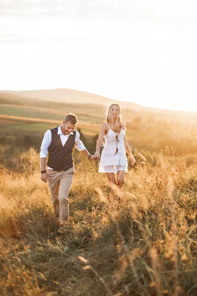 Bel homme élégant en costume rustique et jolie femme boho en robe marchant dans le champ avec des balles de paille, tenant la main et souriant. Soirée d'été, coucher de soleil — Photo