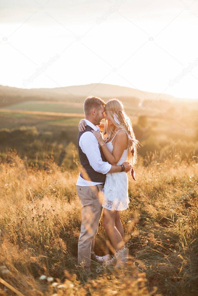 Man and woman in stylish boho casual clothes, posing on the background of mountains, field at sunset. Stylish hipster couple hugging