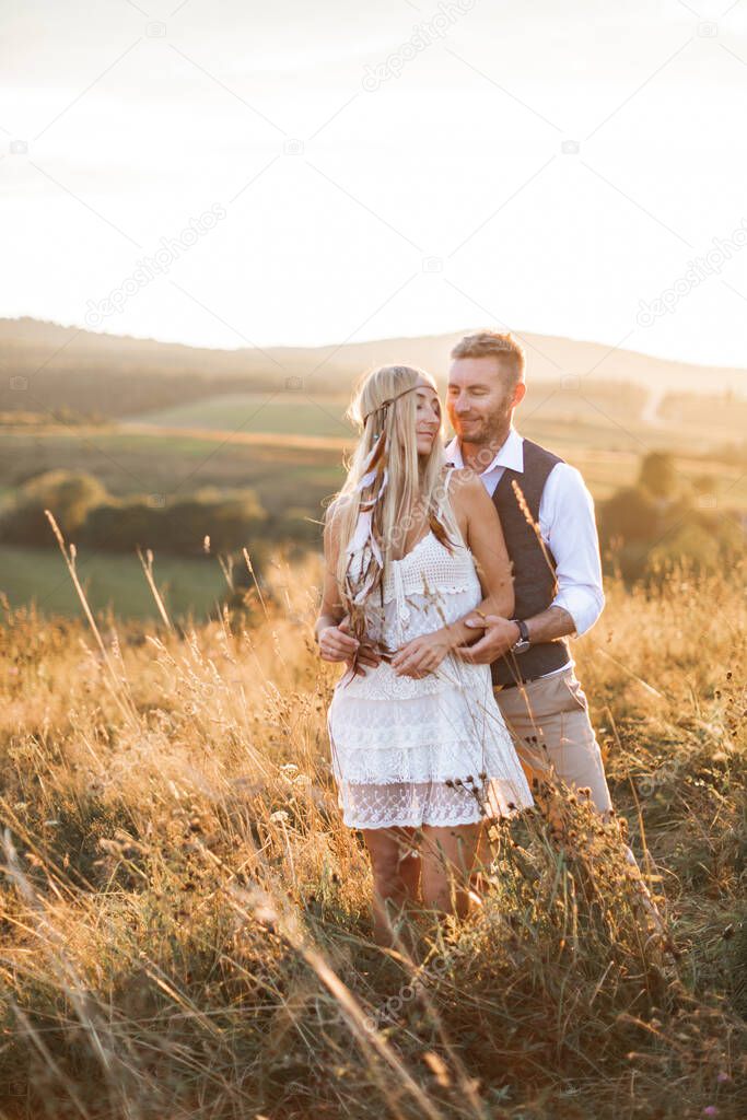 Handsome man hugs his woman standing in the field with straw grass and flowers at summer sunset, outdoors. Wedding, love story in rustic boho style