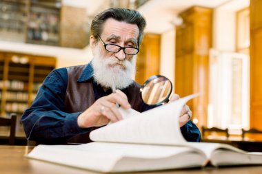 Focused elderly stylish man teacher, university professor, in eyeglasses sitting in old library and turning book pages while looking through the magnifying glass