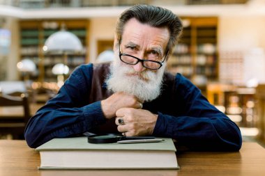 Gözlüklü, gülümseyen beyaz, yaşlı, sakallı bir adamın portresini yakından çek. Elinde kitap, büyüteç ve kalemle antik kütüphanenin arka planında oturuyor.